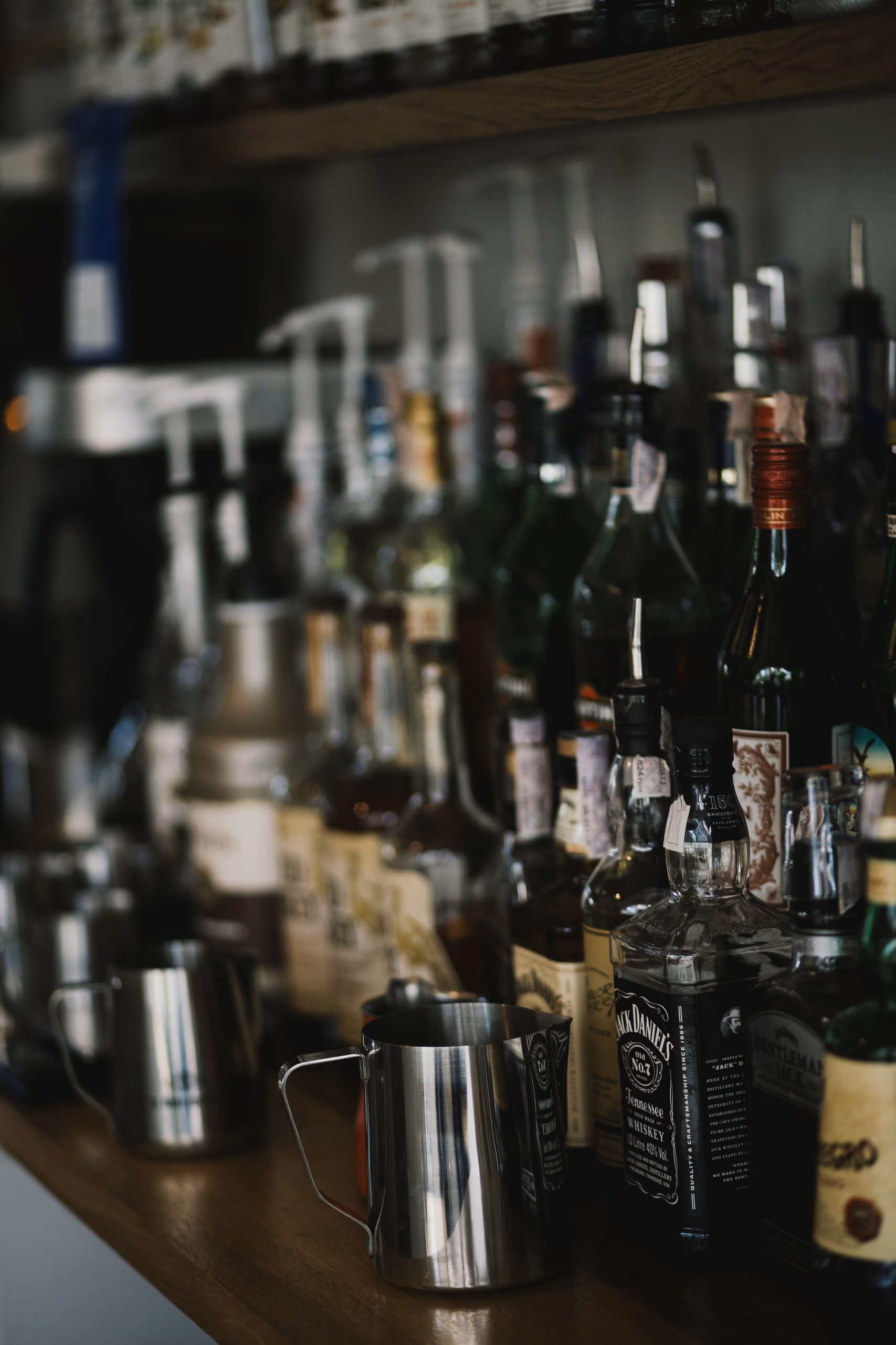 Bottles along a dim bar shelf, a few of them sharp and the rest dissolving into the dark.