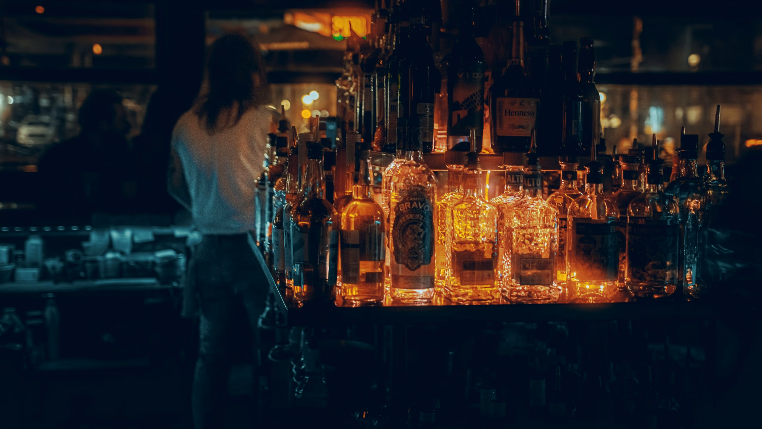 A bar counter at night, bottles lit from below in amber light, one person leaning at the far end.