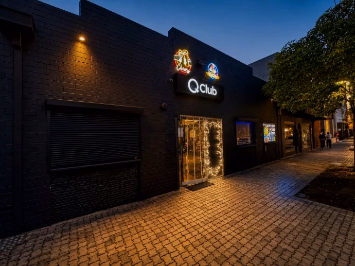 Q Club at dusk: a black painted brick frontage with a lit Q Club sign and two neon signs above a glass door strung with fairy lights, a roller shutter alongside, and a brick-paved footpath running past shopfronts into the street.