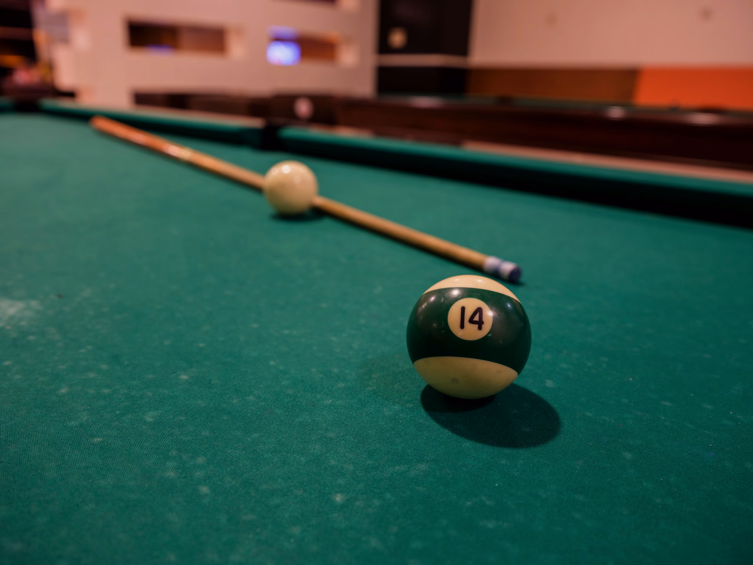 A striped fourteen ball and the tip of a cue on green cloth, the rail glowing warm in the lamp light behind.