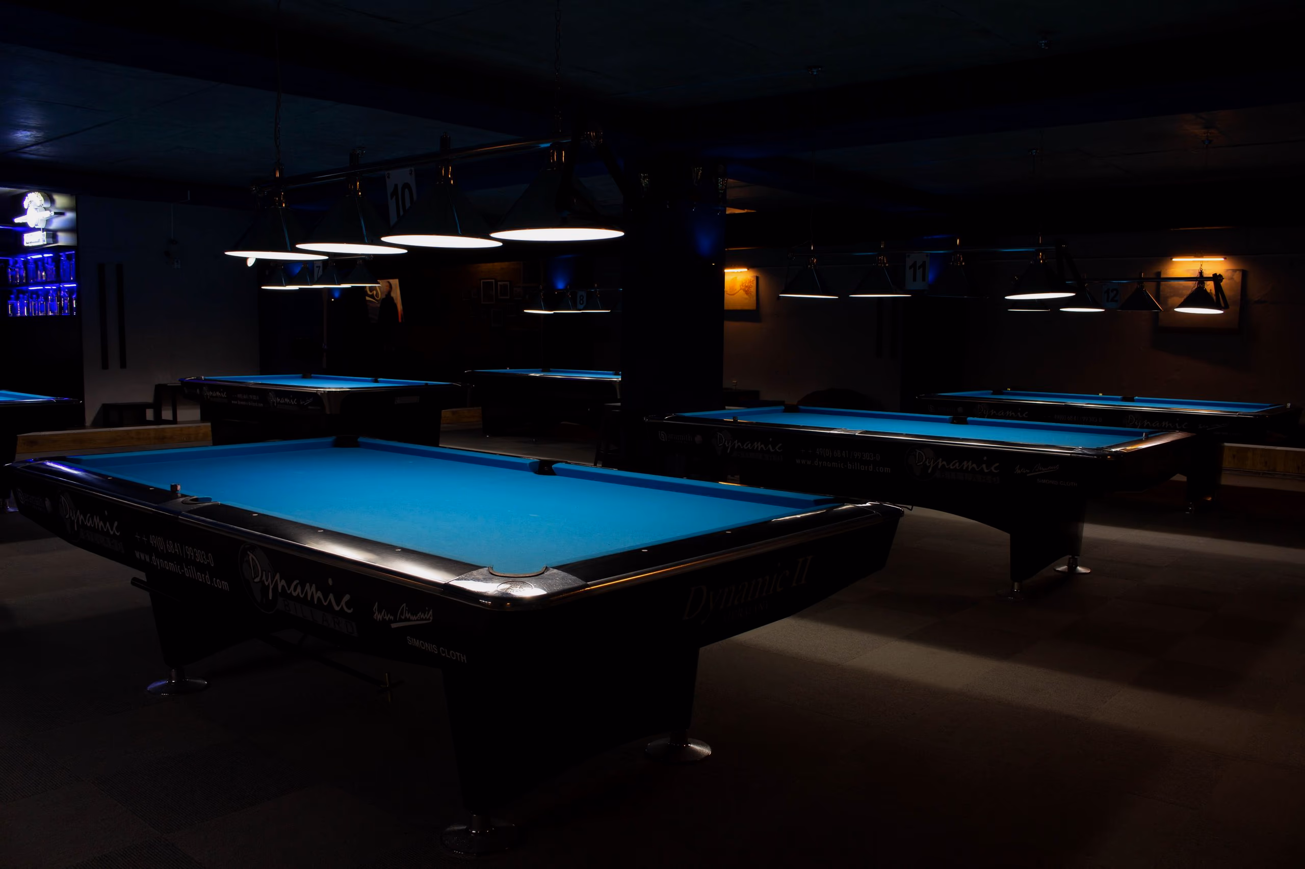 A row of empty blue-cloth pool tables under low lamps in a dim lounge.