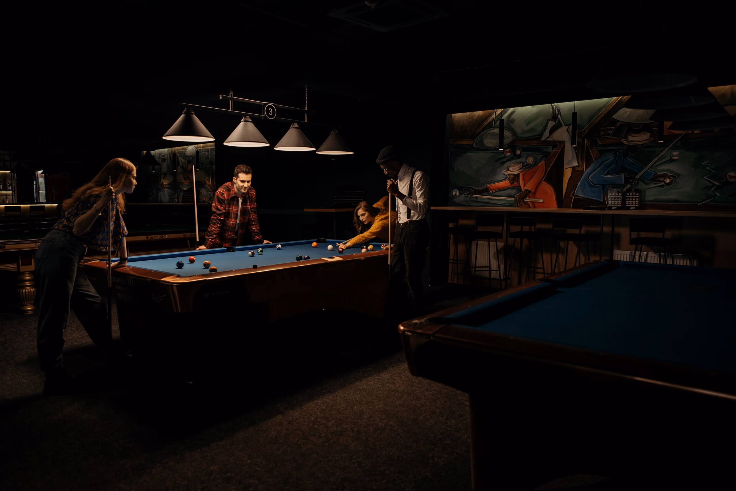Four people around a blue pool table under cone lamps in a dark lounge, one of them down on a shot.