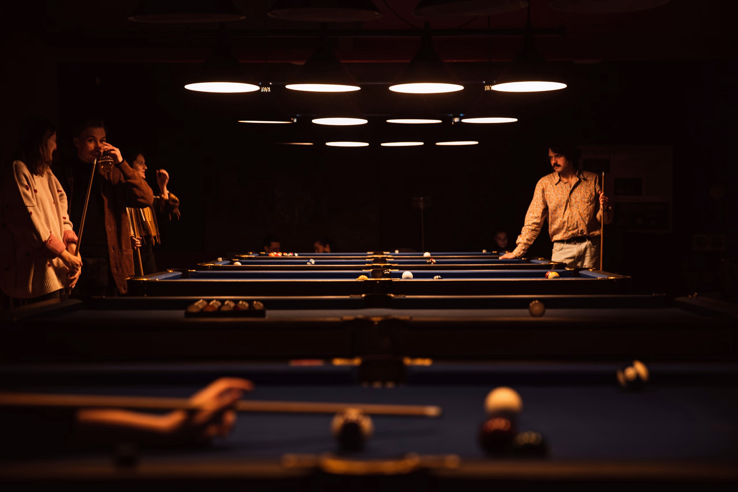 A long row of pool tables under a grid of round lamps in a dark hall, players waiting along both sides and a cue lining up a shot in the foreground.