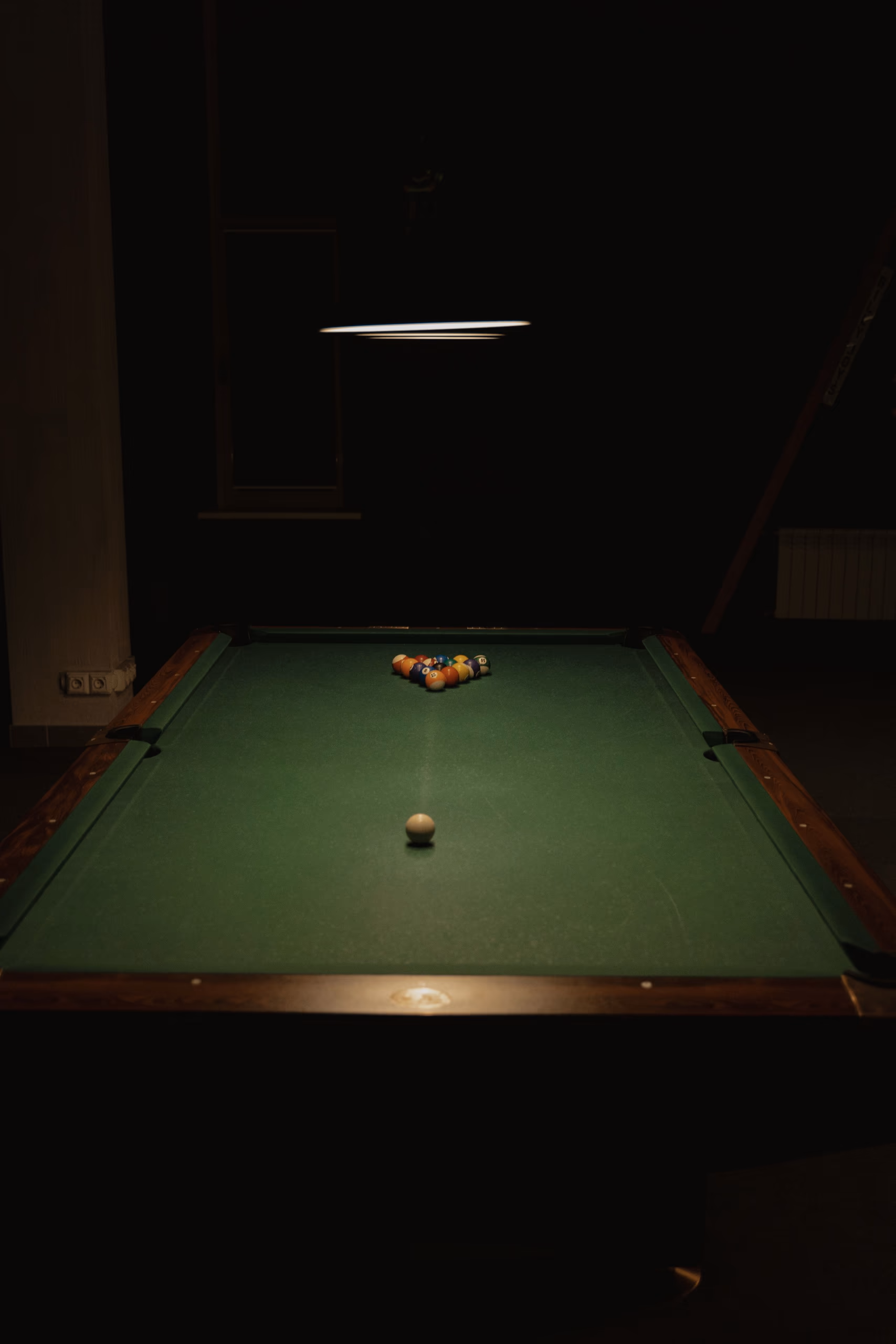 A green-cloth table with a racked triangle of balls, lit by a single lamp in an otherwise dark room.