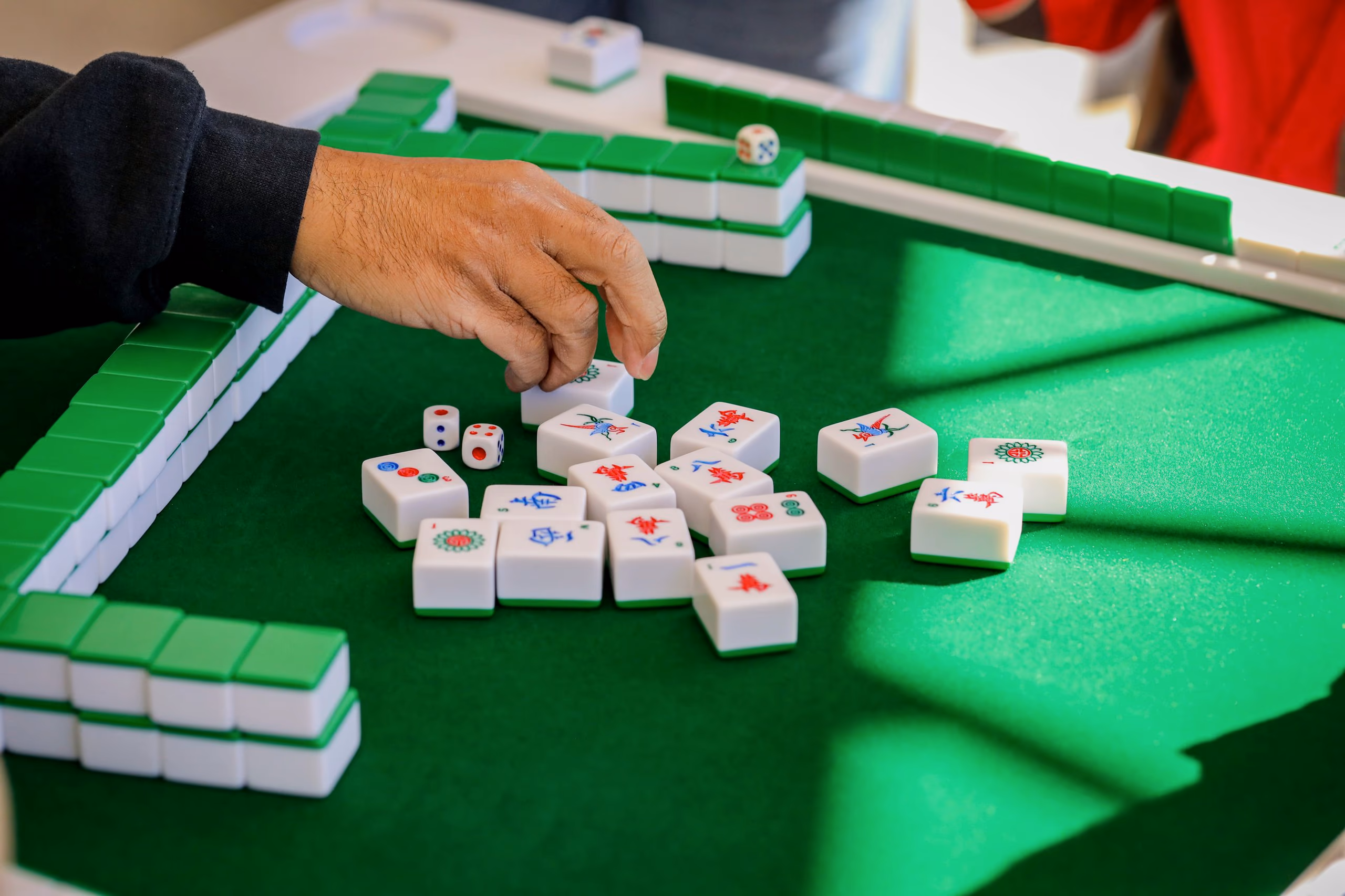 A hand turning a Mahjong tile face-up on green cloth, walls of tiles stacked along the edge of the table.
