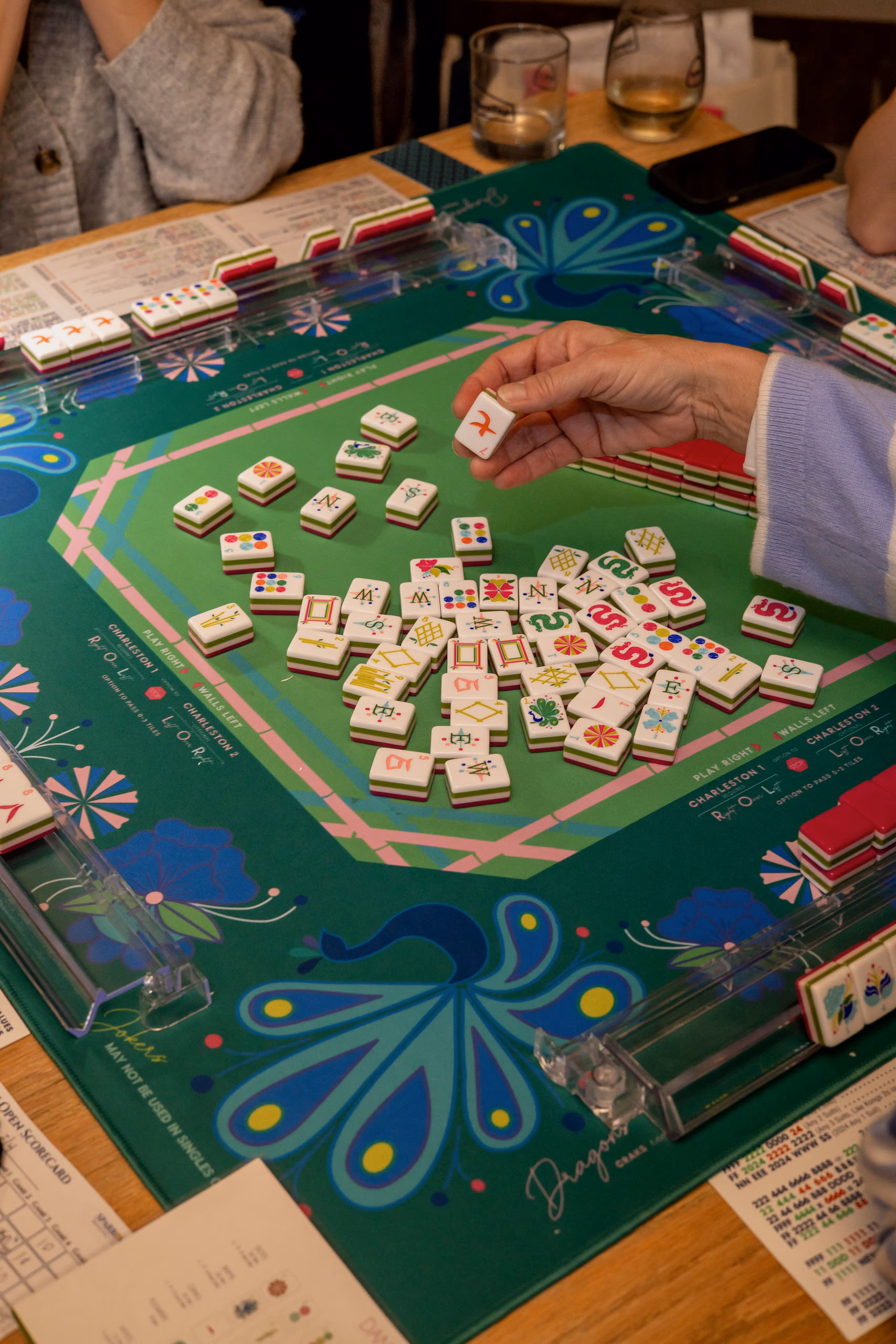 A hand placing a Mahjong tile on a green table printed with a peacock design, tiles scattered mid-game.