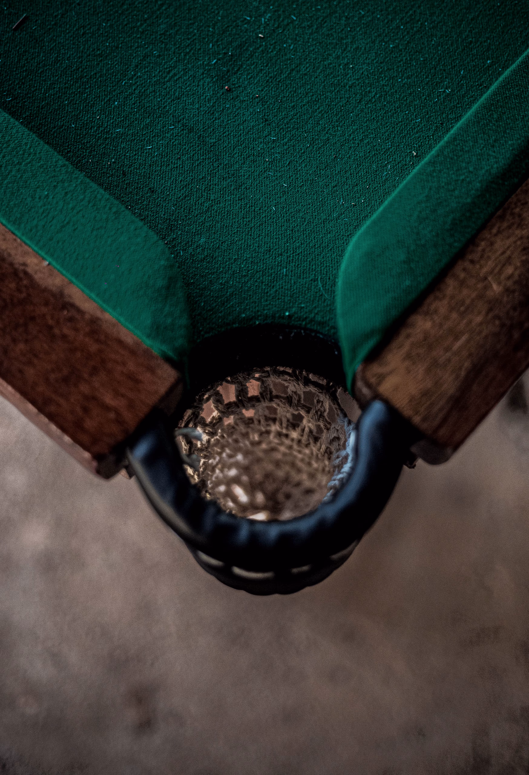 The corner pocket of a billiards table in close-up: green baize, a dark timber rail and the net beneath.