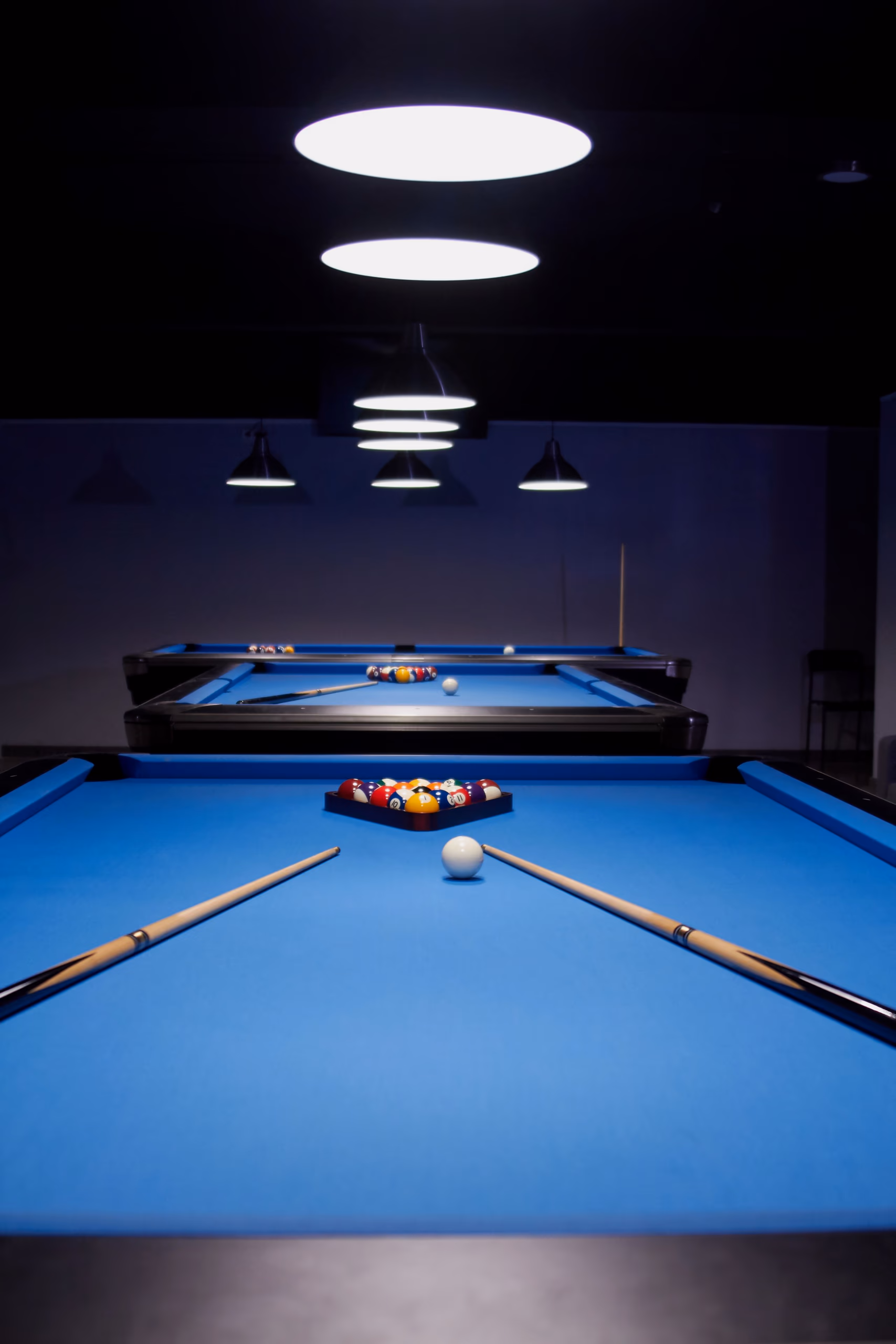 Bright blue pool tables under pendant lamps, two cues laid across the cloth beside a racked set of balls.