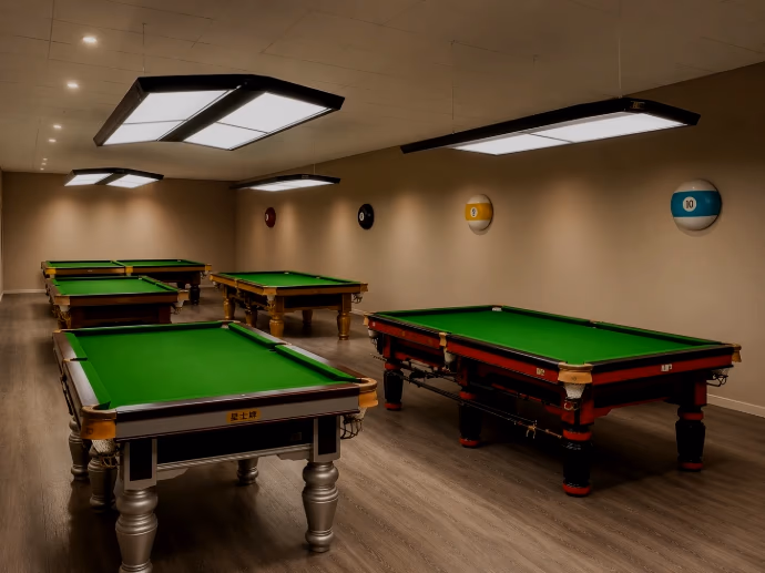 The playing room at Q Club: four billiards tables with green cloth under flat white ceiling lights, three framed in pale timber and one in red, standing on a timber floor, with large coloured billiard balls mounted along the wall behind.