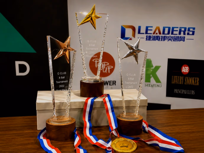 Three star-topped Q Club 8-Ball Tournament trophies and gold medals on striped ribbons, arranged in front of a wall of event sponsor logos.